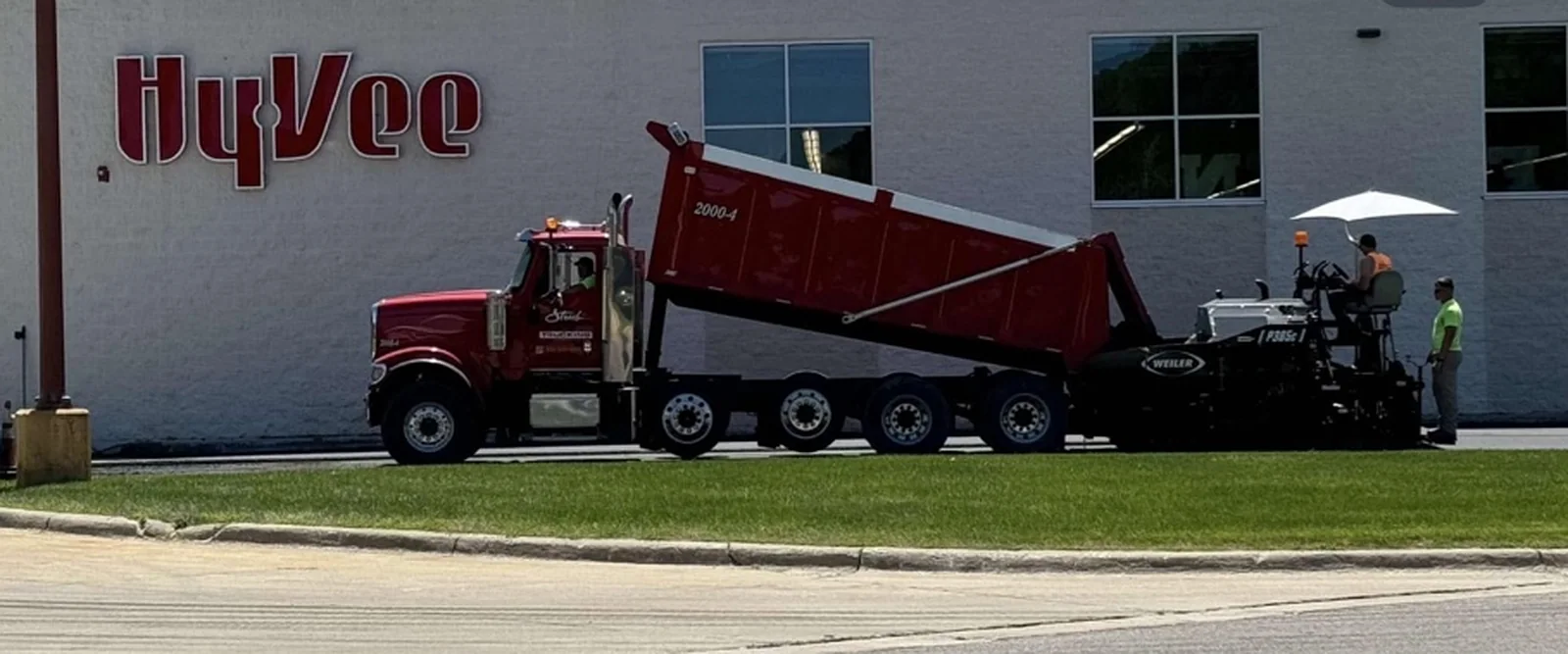 Strub dump truck with its box raised feeding hot asphalt into a Weiler paver at a grocery store