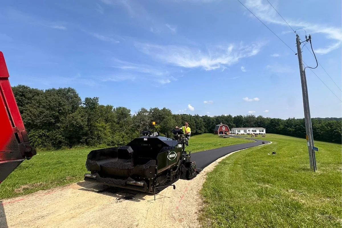 Weiler paver laying hot mix on a new rural driveway running toward a white house and red barn